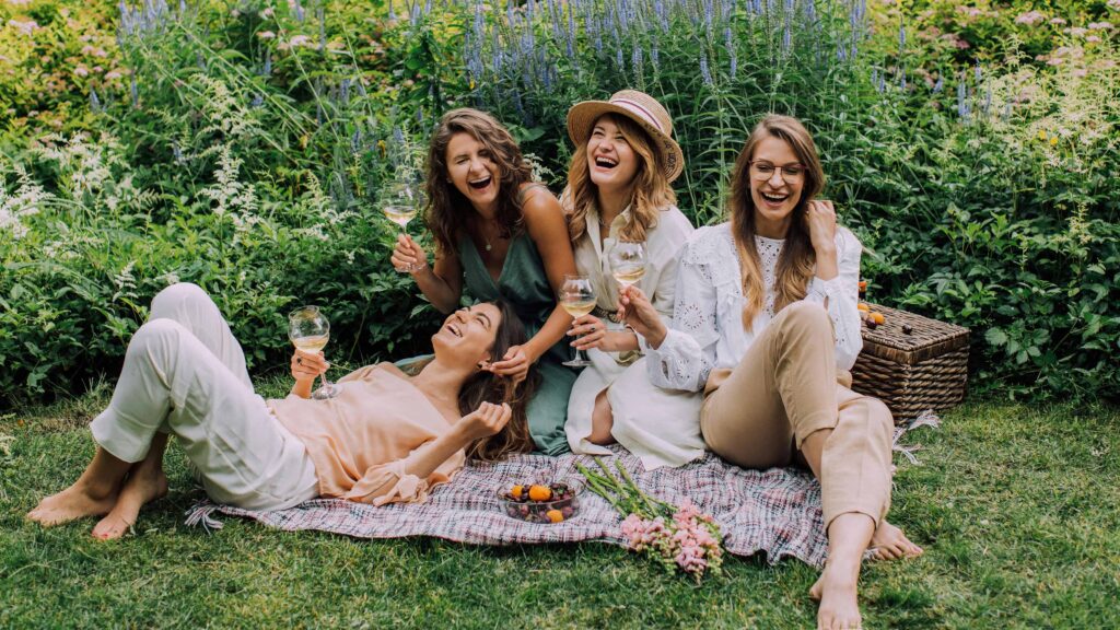 A group of girlfriends share a laugh at an outdoor picnic in a park. From the article: How attachment styles show up in friendships