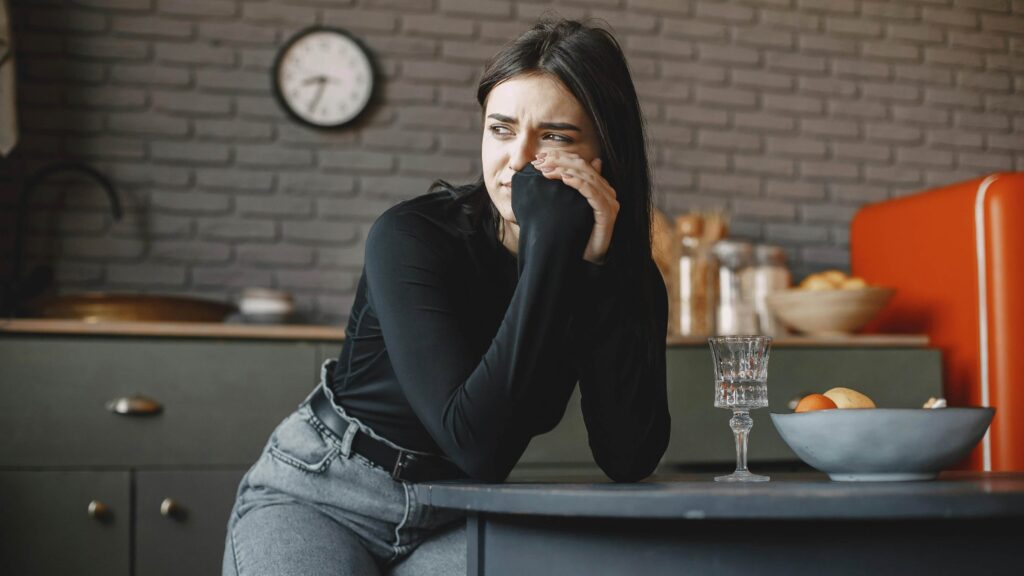 A woman sits anxiously in her kitchen. From the article: "What someone with anxious attachment needs"