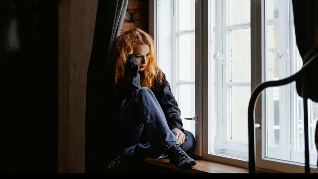 A worried woman sits next to a window. From the article: How to heal from abandonment wounds.