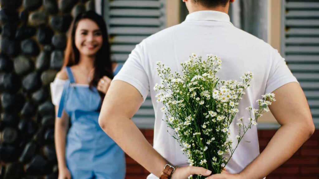 A man holds some daisies behind his back to give to the girl standing in front of him. From the article: How attachment styles affect early dating