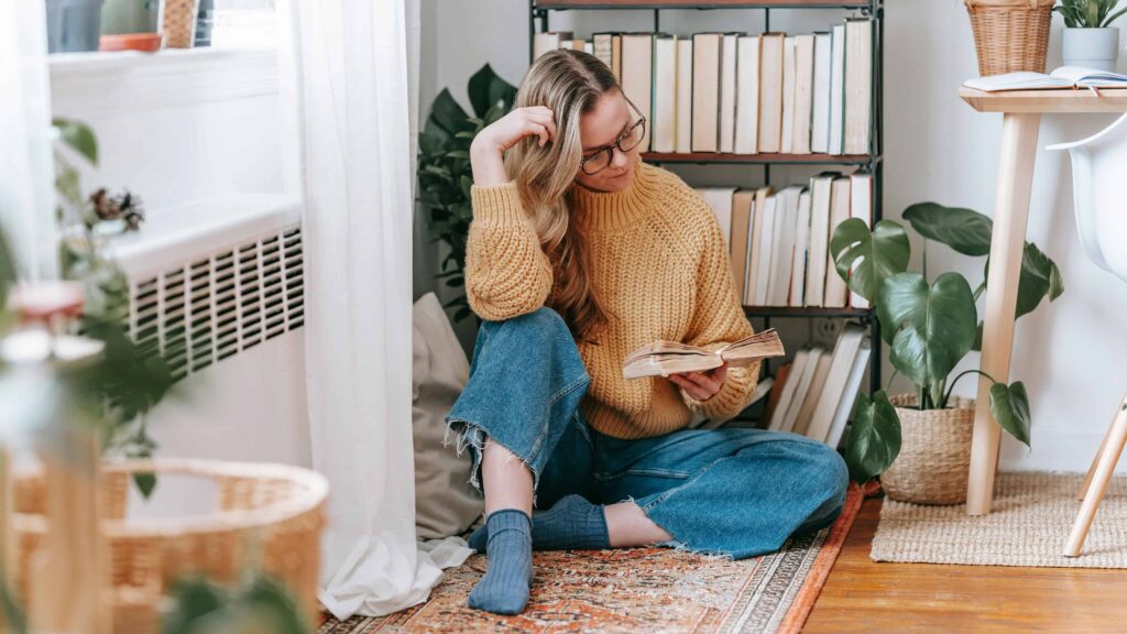 A young woman reads a book in the living room. From the article "books on people pleasing"